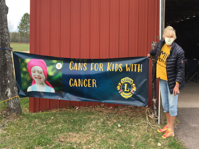 Female Lions Club member standing next to a banner that reads 'Cans for Kids with Cancer'